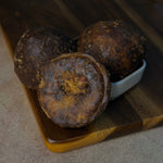 Three dark brown, round, textured smoked karupatti (palm jaggery) , with one cut open to show the interior, arranged on a wooden cutting board next to a small white dish.